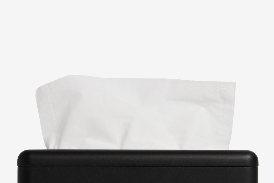 A close-up of a white tissue paper partially pulled from a matte black tissue box dispenser against a clean white background.