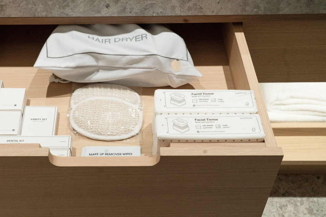 A wooden drawer organizer containing a white hair dryer pouch, a round mesh sponge, Fox Fold facial tissues, and other white toiletry items, photographed in a real interior setting.