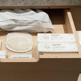 A wooden drawer organizer containing a white hair dryer pouch, a round mesh sponge, Fox Fold facial tissues, and other white toiletry items, photographed in a real interior setting.