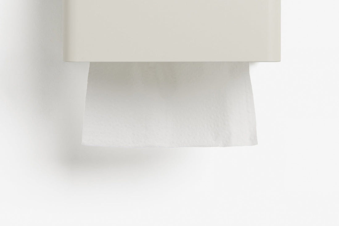 A close-up of a white wall-mounted tissue dispenser with a white tissue paper partially pulled down, set against a clean white background.