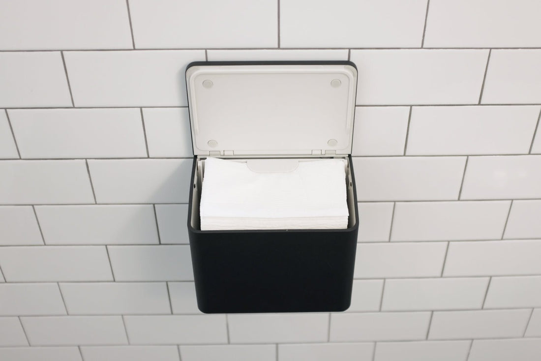 A black wall-mounted tissue dispenser with an open white lid, holding white tissues, mounted on a white subway tile wall.