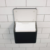 A black wall-mounted tissue dispenser with an open white lid, holding white tissues, mounted on a white subway tile wall.